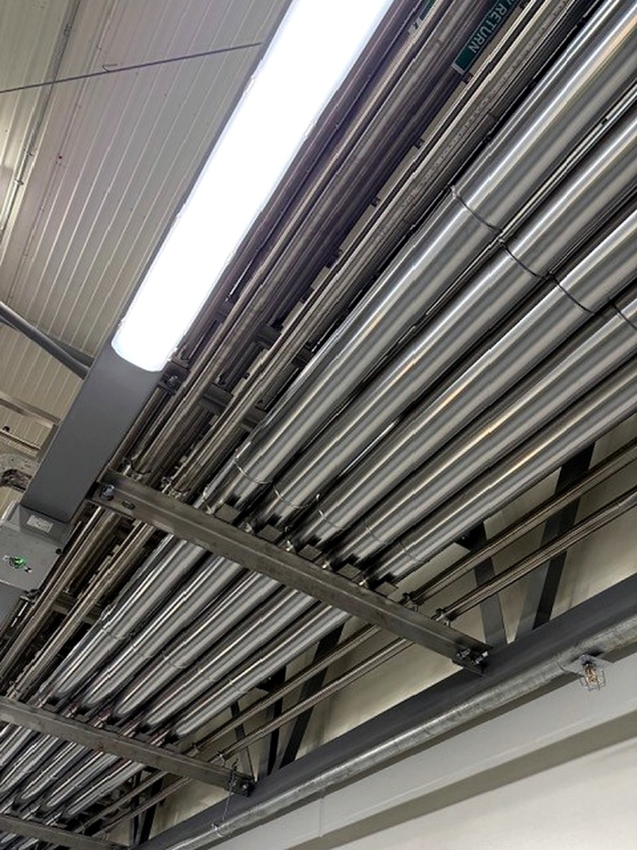 Multiple parallel metal-clad overhead pipe runs viewed from below in an industrial facility